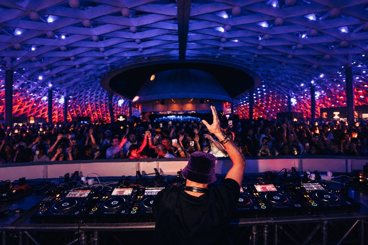 DJ raising a hand to the crowd beneath the blue and purple illuminated gridshell
